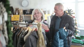 Elderly couple stands in a clothing store against the background of a Christmas tree and chooses jackets and coats for the winter. Pensioners at the New Years sale of outerwear. High quality 4k - Powered by Shutterstock - Get 15% off with code: PIKWIZARD15