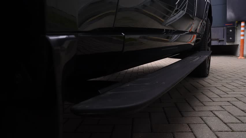 A low angle close up tracks an SUV step deploying at a loading bay with herringbone brick, an orange striped bollard, and a dock unit in soft daylight.