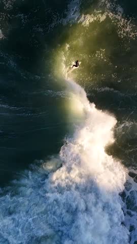 A scenic aerial vertical footage of the beautiful sunlight at sunset illuminating surfer as he riding a wave in Sydney, Australia
