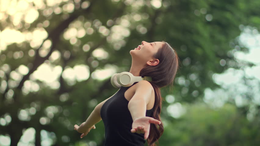 Carefree young Asian woman with arms outstretched enjoying fresh morning air in park at sunrise, wearing headphones and closing eyes, practicing mindfulness, relaxation, peaceful natural surroundings.