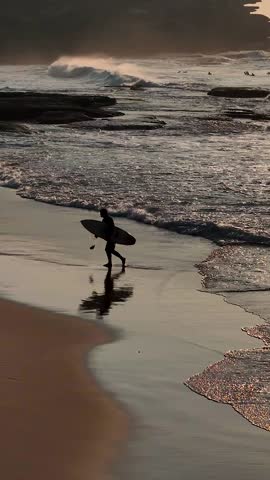 A drone vertical slow-motion of a silhouetted surfer heading back in after a surf and the foamy waves behind them at sunset