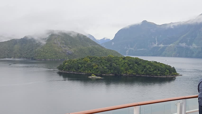 Viewing scenic landscape of Milford Sound in New Zealand exploring the fjord from cruise ship, showing small forested island with mist shrouded mountains and waterfalls