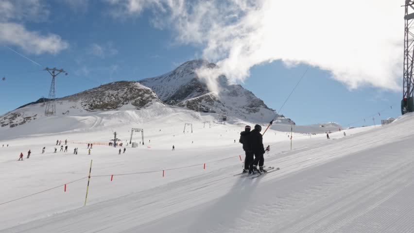 A slow-motion of skiers under aerial cableway surrounded by mountains and blue sky with white clouds in Austria