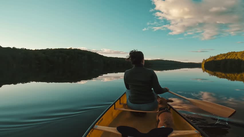 A slow-motion, back shot of a woman gracefully paddles her canoe across the serene waters of Algonquin Provincial Park in Ontario, Canada at sunset