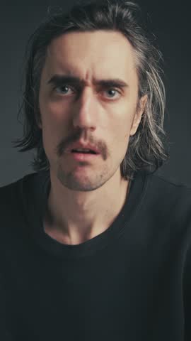 young attractive funny comic European man with mustache in a black shirt and long hair shows a silly confused expression in close-up shots