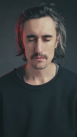 Playfully peeking, a young attractive European man with a mustache and long hair in a black sweatshirt humorously squints and winks at the camera in a dimly lit atmosphere.
