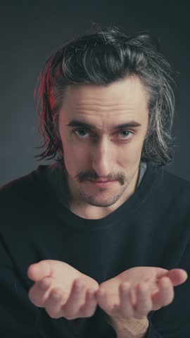 Begging: A humorous young attractive European man with a mustache and long hair, playfully looking into the camera with a pleading gesture, dressed in a black shirt.