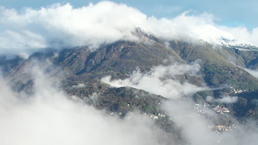Aerial view from a drone showcasing the stunning Alps in Italy, featuring majestic mountains partially covered by clouds amidst a serene landscape.
