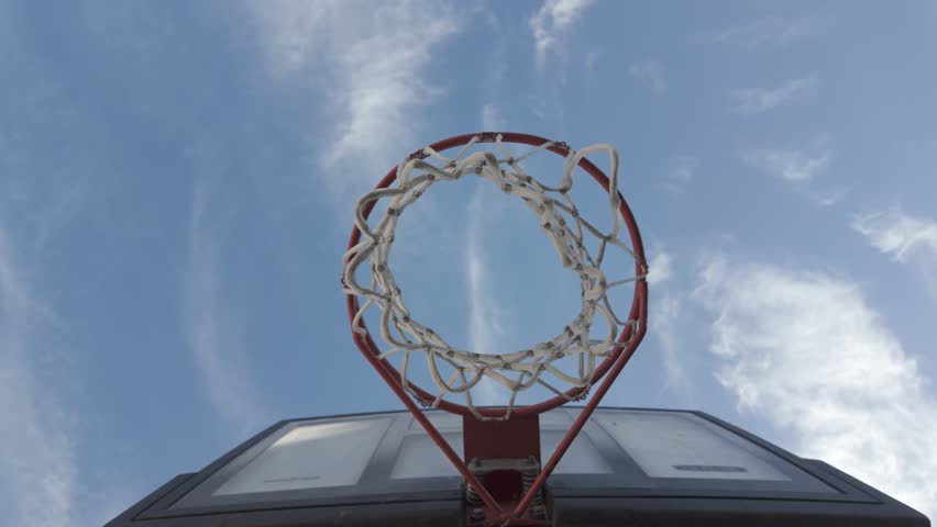 A slow-motion low angle view of a basketball hoop under blue sky with partial clouds, on a sunny day