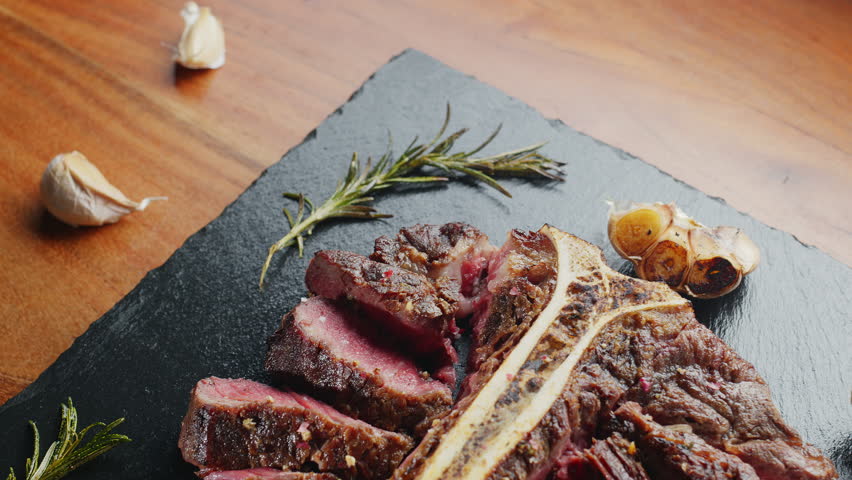 Sliced medium-rare T-bone steak with herbs and garlic on the serving slate board close-up. Top view.