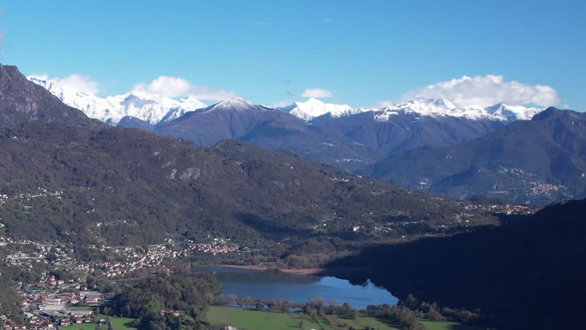 This aerial view captures the breathtaking beauty of the Italian Alps, showcasing snow-capped peaks over a peaceful lake surrounded by lush greenery.