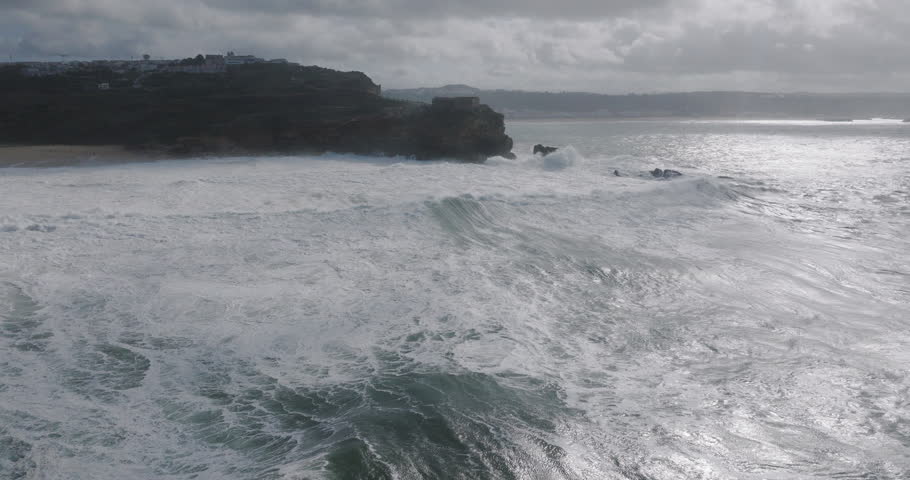 Aerial drone shot of big waves breaking on rocks and cliffs on a day with giant waves in Nazare, Portugal, Europe. Farol da Nazaré lighthouse visible. Shot in ProRes 422 HQ