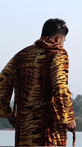 A flipped vertical shot of Indian male model wearing a tiger print robe and relaxing by the water
