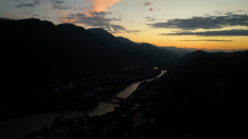 A dark, moody aerial view of a valley town beside a winding river, captured at sunset with strong orange and purple tones behind the mountain silhouette.