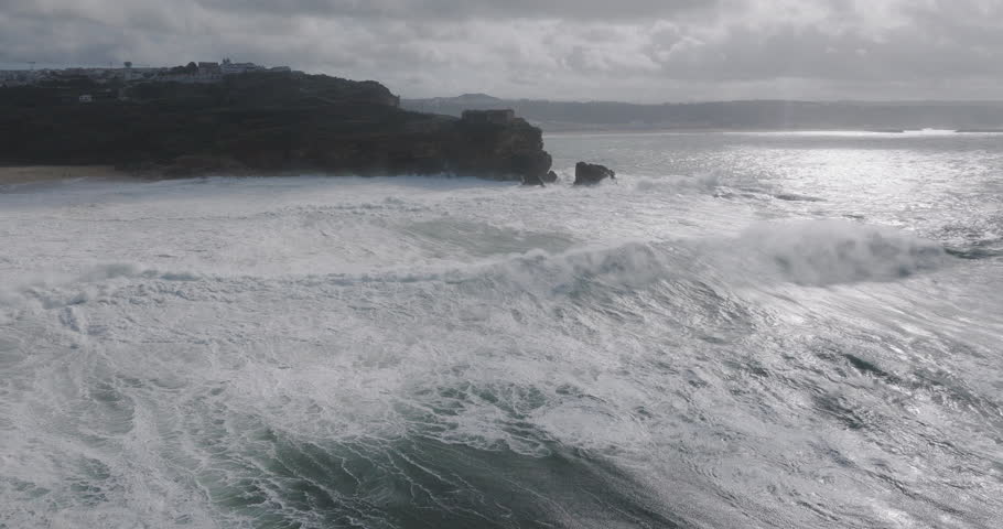 Aerial drone shot of big waves breaking on rocks and cliffs on a day with giant waves in Nazare, Portugal, Europe. Farol da Nazaré lighthouse visible. Shot in ProRes 422 HQ