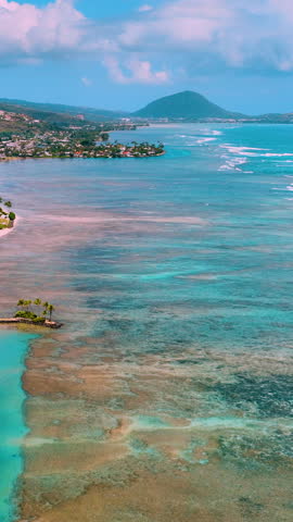Shallow coral reefs and vibrant blue waters near shoreline with buildings and mountain backdrop. Aerial view, Hawaii. Vertical video.