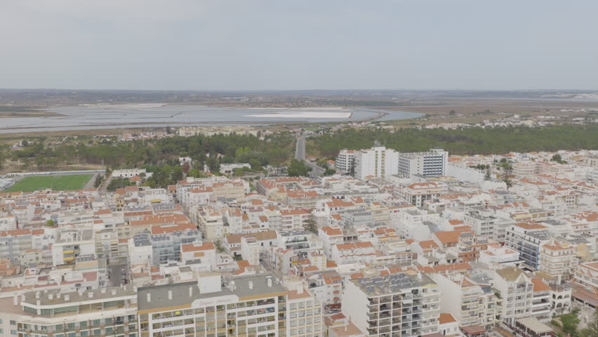 High-altitude drone flies forward over Monte Gordo rooftops toward the salt marshes and lagoon area, revealing urban patterns, green zones, and water basins.