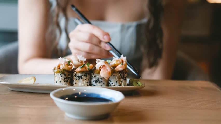 A beautiful girl eats Japanese sushi rolls with chopsticks, smiling with pleasure