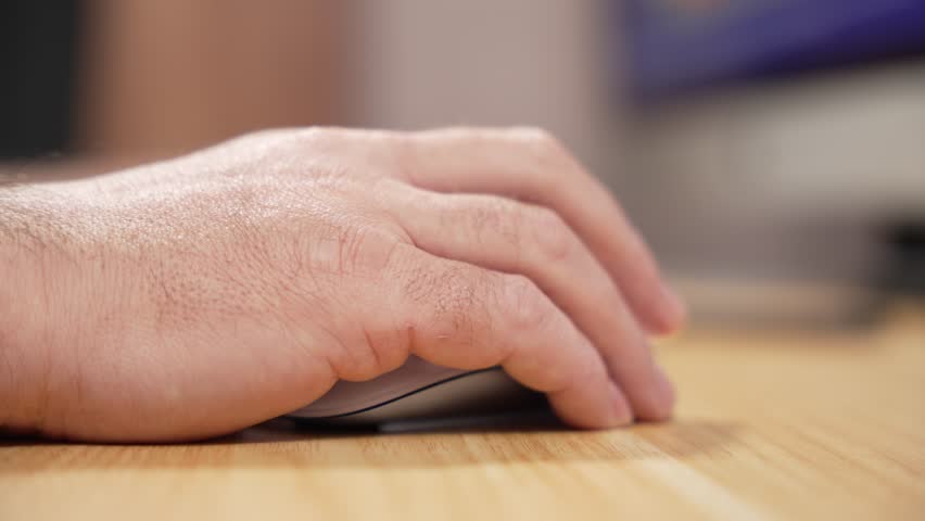 Close-up video of a mans hand resting and clicking on a computer mouse on a wooden desk. Subtle movement suggests focused office or home office work.