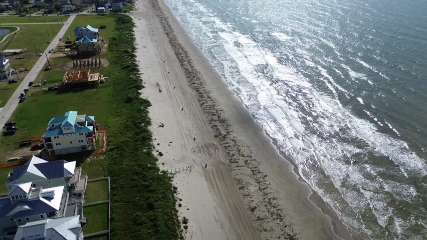 Beachfront homes under construction near Sunny Beach, Galveston. Tire tracks, dune vegetation, and shimmering waves highlight the balance of natural shoreline and elevated residential expansion. USA