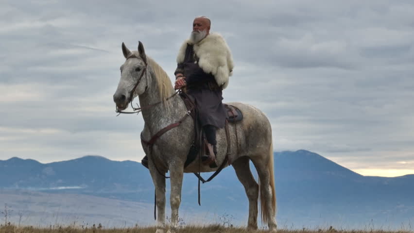 Aerial drone establishing or tracking shot of Bearded warrior in fur cloak riding a white horse across open landscape at sunrise, mountains in the background. Historical, rugged, adventurous scene