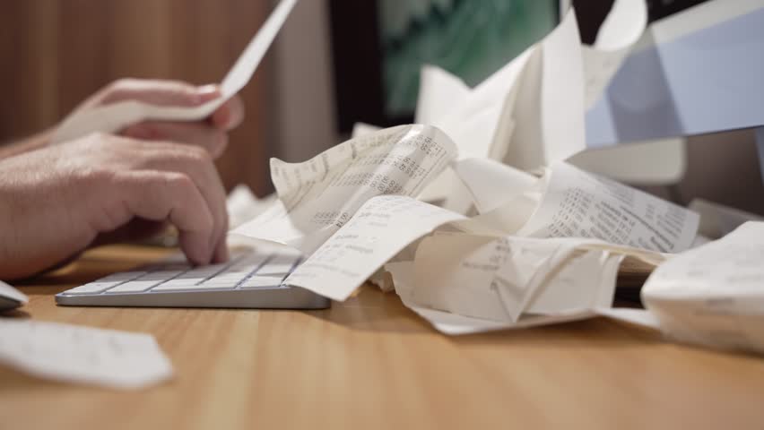 Man types on a keyboard while surrounded by crumpled paper receipts in this video. The clip illustrates chaotic bookkeeping or stressful tax season.