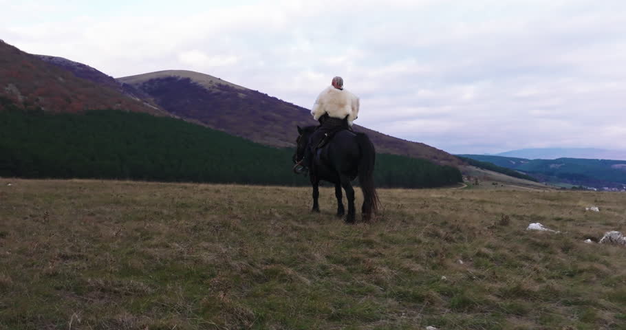 Cinematic aerial drone shot of a Viking warrior on a large black horse surveys a dramatic landscape during sunset, wearing a fur cloak concept of Ancient exploration, adventure, and the spirit of the