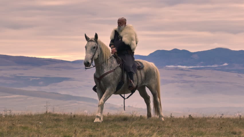 Aerial drone establishing or tracking shot of Bearded warrior in fur cloak riding a white horse across open landscape at sunrise, mountains in the background. Historical, rugged, adventurous scene
