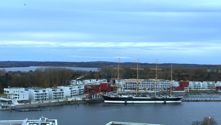 a panoramic view over the waterfront and marina area. A large sailing ship Passat is moored at the pier, standing out prominently against the modern white and red residential buildings nearby.