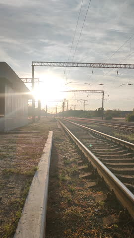 Male workers in uniform orange vests with reflective strip walking along railway track near railway station. Railway station platform. Rails sleepers. Shining sun at dawn. Railway Vertical