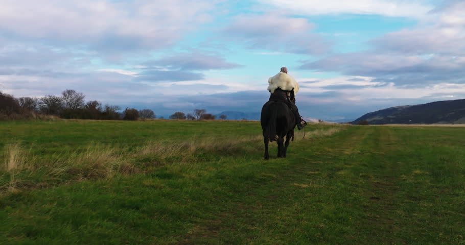 Cinematic aerial drone shot of a Viking warrior on a large black horse surveys a dramatic landscape during sunset, wearing a fur cloak concept of Ancient exploration, adventure, and the spirit of the