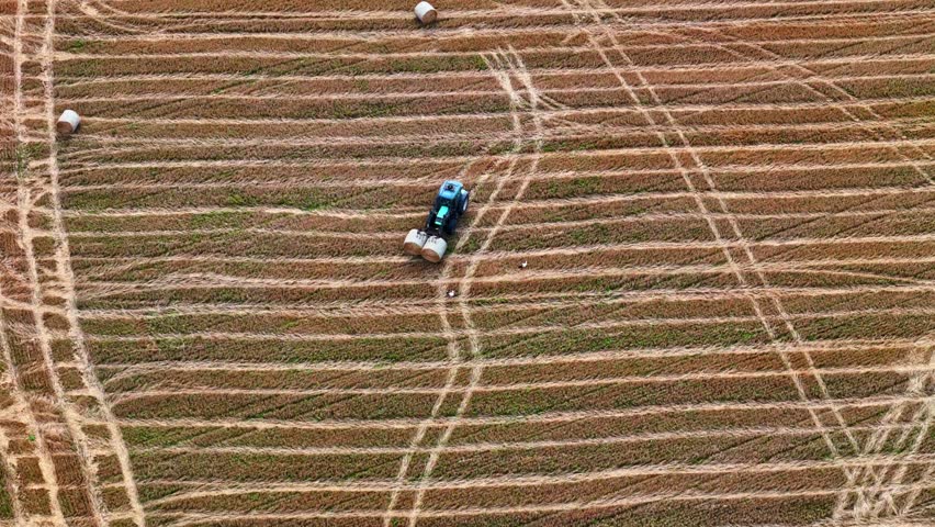 Blue tractor baling hay on patterned farmland with visible tire tracks