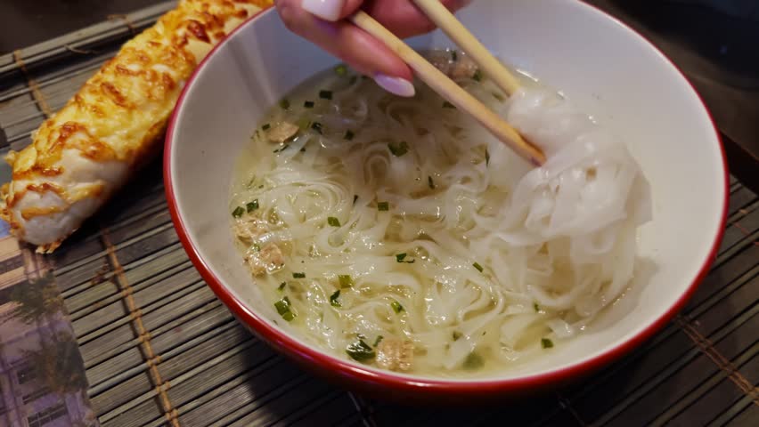 Eating Rice Noodles In Clear Broth With Chopsticks, Delicious Asian Soup Meal, Hand Holding Chopsticks Over Bowl. Authentic Southeast Asian Flavors. Vietnamese Pho Or Ramen. Traditional Asian Cuisine