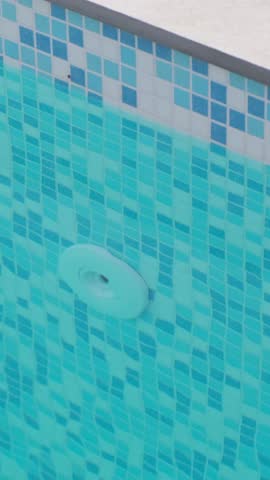 Fragment of a swimming pool wall covered with blue mosaic tiles, featuring a visible water inlet nozzle. Clear water creates ripples that form a pleasant surface pattern

