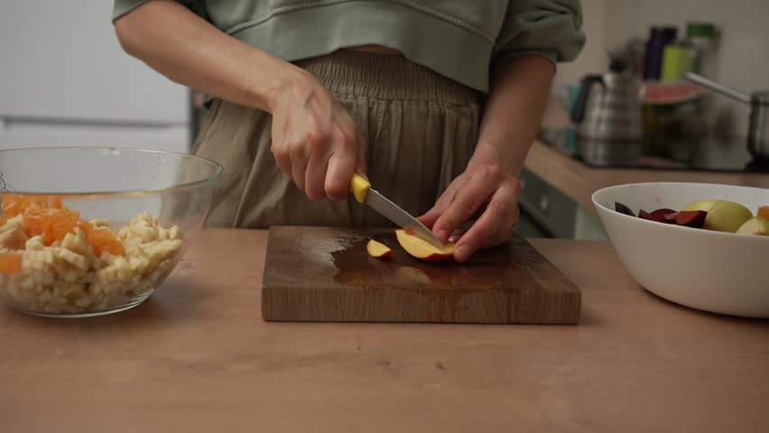 Close-up of a woman slicing a fresh peach on a wooden cutting board to prepare a healthy and delicious fruit salad in a modern kitchen, promoting a wholesome and nutritious lifestyle