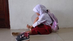 An Indonesian Muslim elementary student sits on the floor putting on socks and sneakers before leaving for school. Focus on preparation, education, and back-to-school routines. - Powered by Shutterstock - Get 15% off with code: PIKWIZARD15