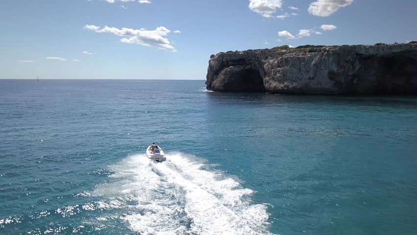 A drone view of a speedboat leaves a wake as it travels across clear blue water, near a large rock formation under a partly cloudy sky.