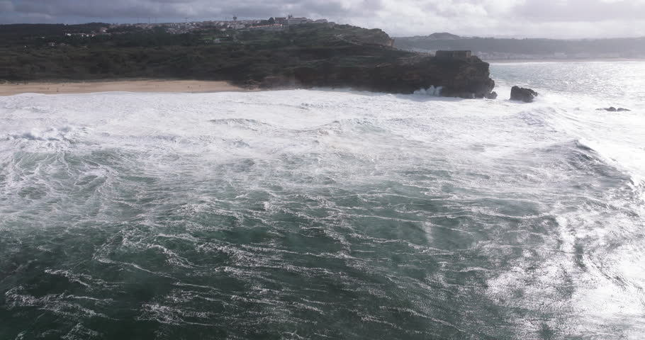 Aerial drone shot of big waves breaking on rocks and cliffs on a day with giant waves in Nazare, Portugal, Europe. Farol da Nazaré lighthouse visible. Shot in ProRes 422 HQ
