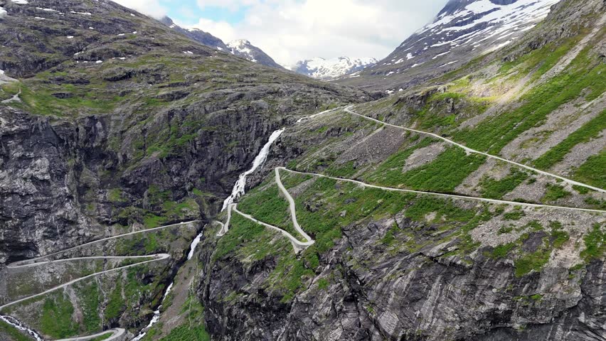 A drone view of dramatic switchbacks on Trollstigen road ascending rocky mountain slopes with patches of snow under a partly cloudy sky