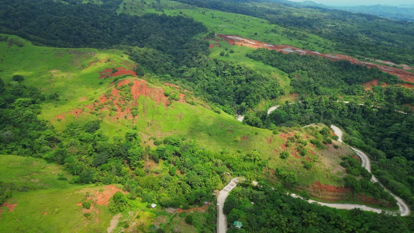 Downward aerial of Quinawan Mountain View in Mariveles, Bataan, highlighting rolling green hills and winding roads with scattered sunray spots illuminating the landscape.