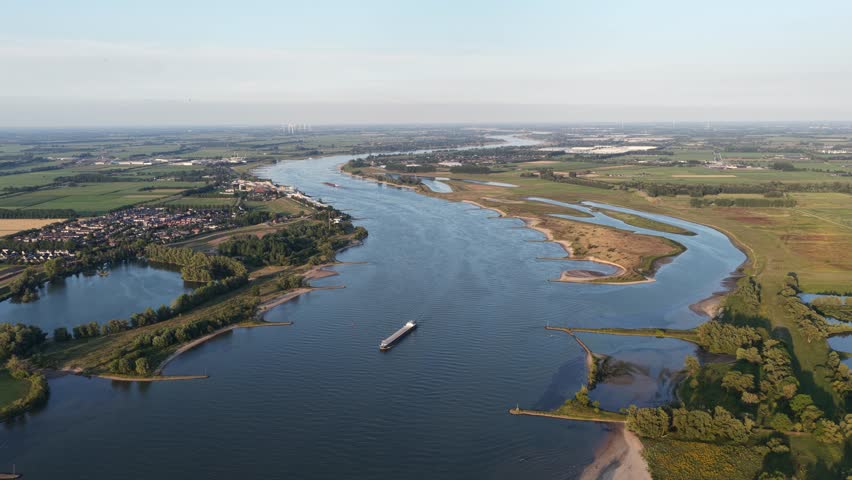 An aerial view of the Waal River at sunset, with a cargo ship navigating vital inland shipping route near a Dutch town and surrounding floodplains