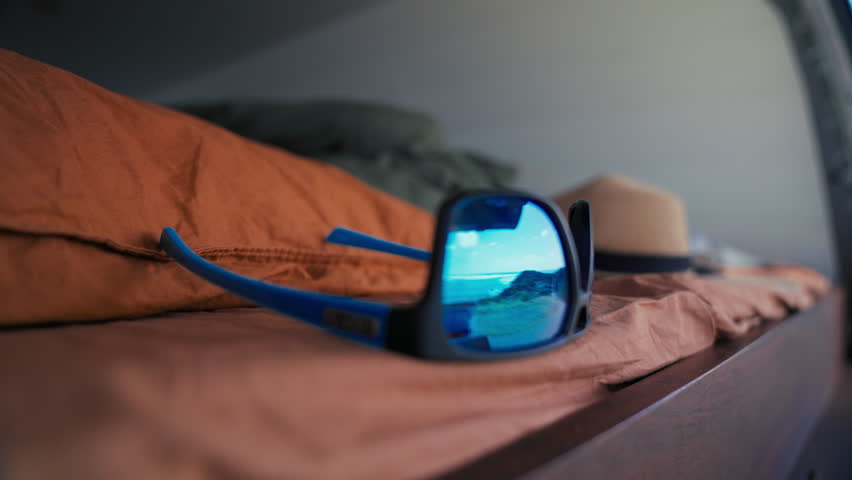 Sunglasses lie on a bed, reflecting a sunny beach scene, with a straw hat nearby in a van