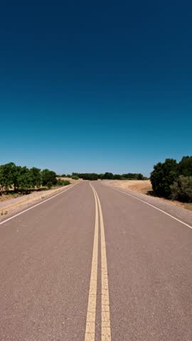 A vertical footage of a straight, two-lane road with a double yellow line stretches into the distance under a clear, deep blue sky. Shrubs on sides