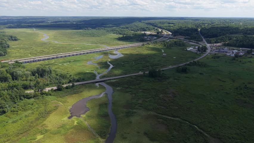 An aerial view of an interstate highway crossing over a vast swampy wetland, surrounded by lush greenery under a partly cloudy sky
