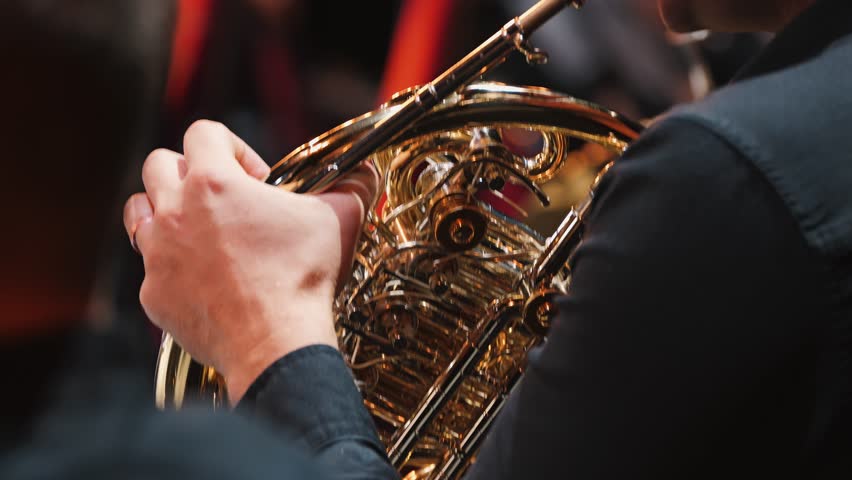 Close-up of the French horn playing. The musician's hand presses the keys of a brass French horn, displaying intricate valves and tubes. The brass section of the orchestra