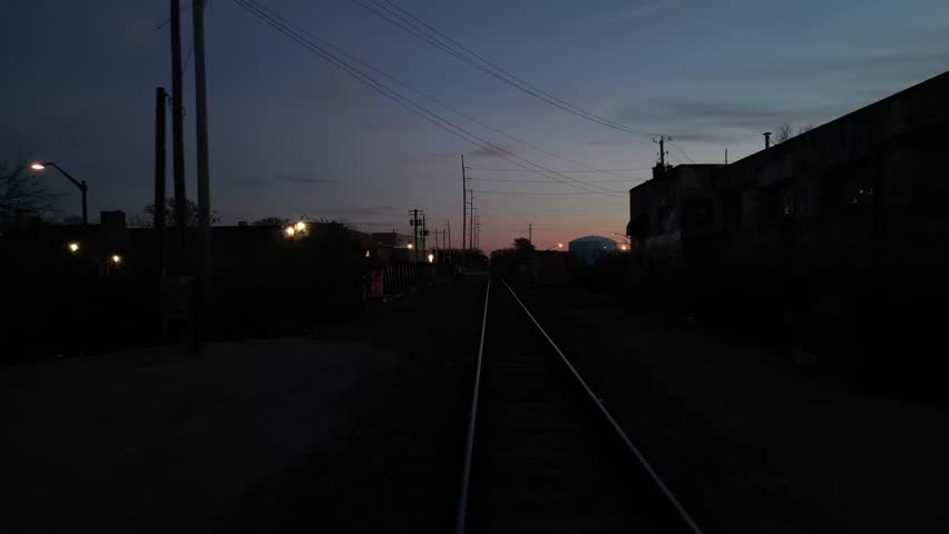 A view down a lonely railway track at twilight, flanked by shadowy buildings and lit by distant streetlights under a colorful, fading sky
