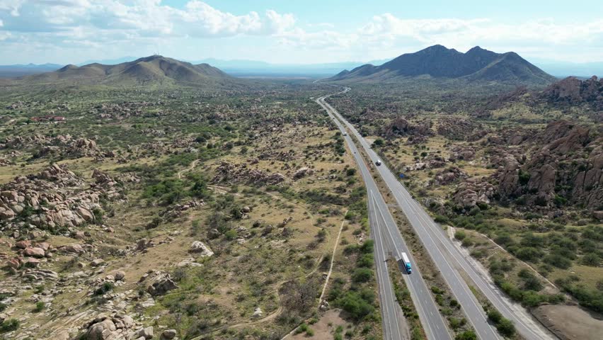 An aerial view shows a multi-lane highway cutting through a vast, arid, rocky desert landscape of Texas Canyon with distant mountains in Arizona