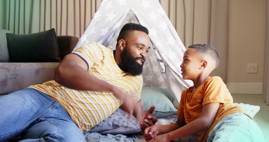 Father and son reclining in teepee, father handing handheld toy and sparking learning, HUD rotating. Parent, child, bonding, interaction, play, tent, cushions