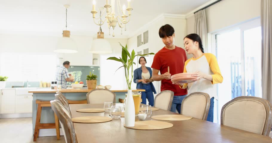 Four adults bringing plates and arranging dining table for food, animated icons floating over hands. Family, kitchen, openplan, teamwork, dishes, serving, placemats