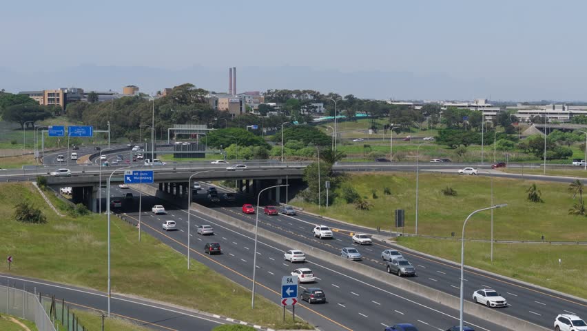 A drone pullback view of cars driving on Observatory Road under a bridge, with greenery surrounding at the daytime in Cape Town, South Africa